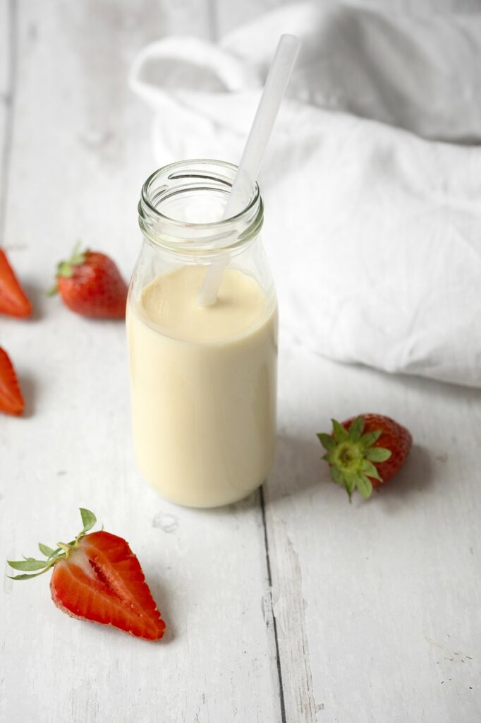 Lait d'avoine maison dans une petite bouteille en verre avec une paille, entourée de fraises fraîches entières et coupées sur un fond en bois blanc.