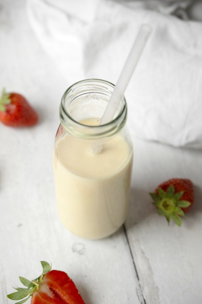 Vue plongeante sur une bouteille en verre remplie de lait d'avoine maison à la texture fluide et crémeuse, avec une paille et des fraises fraîches autour.