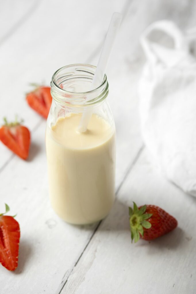 Bouteille de lait d'avoine maison avec une paille posée sur une table en bois blanc, accompagnée de fraises fraîches coupées en deux et d'un torchon blanc.